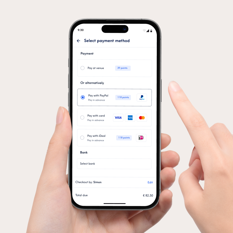 Primer plano de un teléfono inteligente que muestra la interfaz de pago de la aplicación Treatwell para seleccionar el método de pago. Entre las opciones se encuentran el pago en el local o el pago por adelantado con PayPal, tarjeta de crédito o iDeal. Una mano sostiene el teléfono y un dedo señala la pantalla.
