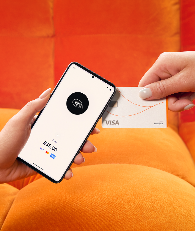 Una mano sostiene un teléfono inteligente que funciona como datáfono, mostrando un icono de pago contactless y un importe total de 35,00 libras, mientras otra mano acerca una tarjeta de crédito Visa blanca para realizar el pago. El fondo es un sofá de color naranja brillante.