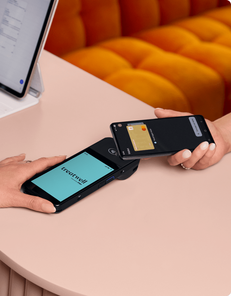 Una persona sostiene un datáfono negro con la pantalla azul claro de la marca "Treatwell", mientras otra persona acerca su teléfono inteligente para realizar un pago contactless a través de una billetera digital. La escena se desarrolla sobre una mesa rosa claro.