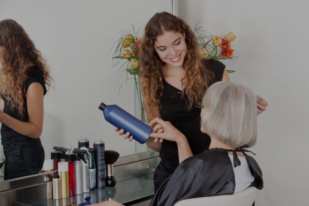 Una joven peluquera sonriente con el pelo rizado sostiene un frasco azul de producto capilar mientras asesora a una mujer mayor de cabello plateado sentada en un sillón de peluquería.
