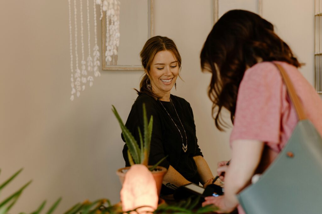 Una dependienta o recepcionista sonriente con el pelo castaño recogido y una camiseta negra mira a una cliente de espaldas mientras utiliza un datáfono sobre el mostrador. En el mostrador hay una planta de aloe vera y una lámpara de sal rosa encendida.