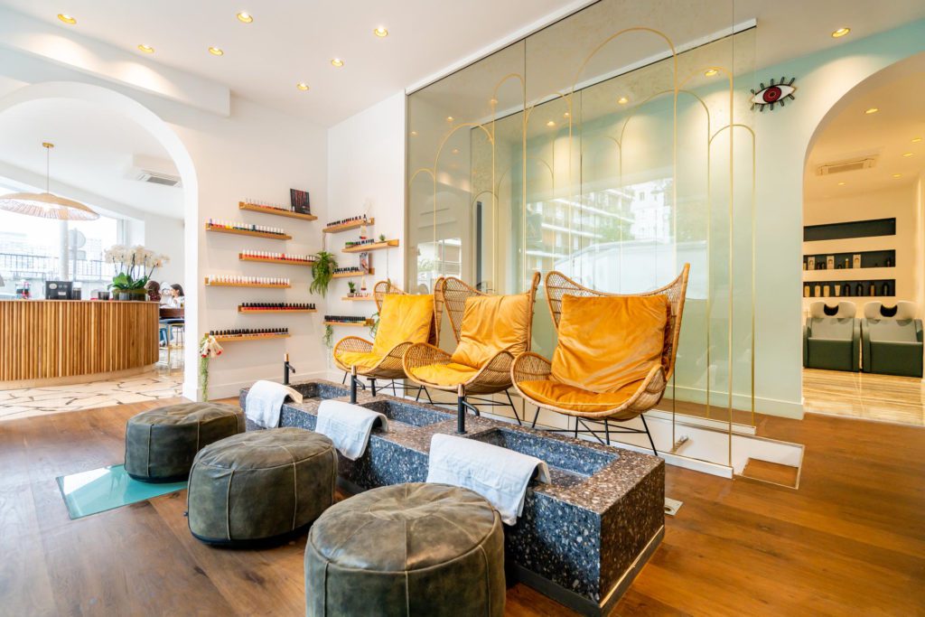 Elegante e iluminado interior de un salón de belleza que cuenta con una zona de pedicura con tres sillones de mimbre color amarillo mostaza, lavabos de piedra con toallas blancas y pufs grises a juego sobre un suelo de madera.