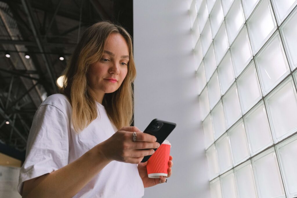 Una toma en contrapicado de una mujer joven con cabello rubio, vestida con una camiseta blanca, mirando hacia su teléfono inteligente negro mientras sostiene un vaso de café para llevar rojo. Está de pie junto a una gran pared de bloques de vidrio.