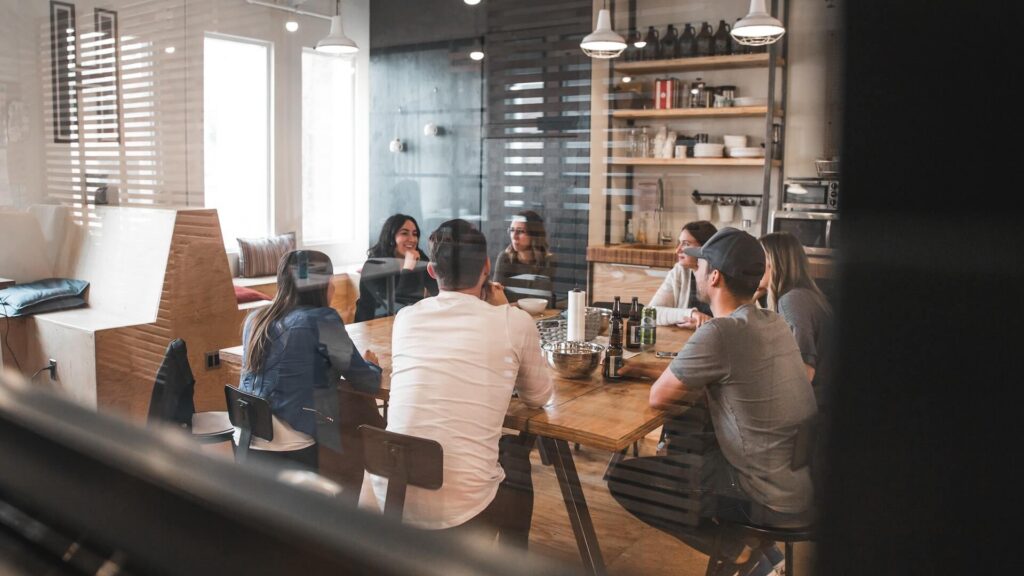 Vista natural a través de un cristal de un grupo de compañeros de trabajo sentados alrededor de una larga mesa de madera en la cocina o comedor de una oficina moderna, conversando y tomando algo.